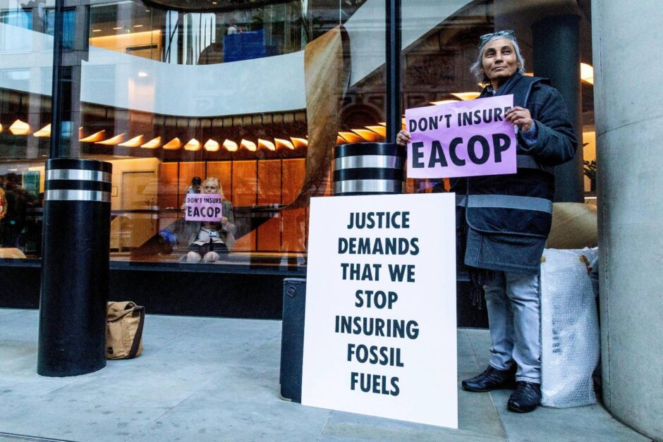 Extinction Rebellion Insure Our Survival protester outside a corporate building, demanding that insurers stop underwriting fossil fuels. The woman holds a sign reading 'DON'T INSURE EACOP' and stands next to a large placard stating 'JUSTICE DEMANDS THAT WE STOP INSURING FOSSIL FUELS'. This protest targets the insurance industry's role in oil and gas projects like the East African Crude Oil Pipeline.