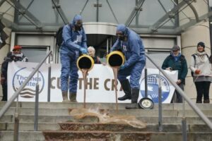 Photograph of a protest, apparently outside of a corporate building, apparently Thames Water. Two figures in blue hazard suits and black gas masks pour a brown substance from barrels down steps. There are two more people holding the banner, one at the side, and a "Cut the Crap" banner with Thames Water and Dirty Water logos